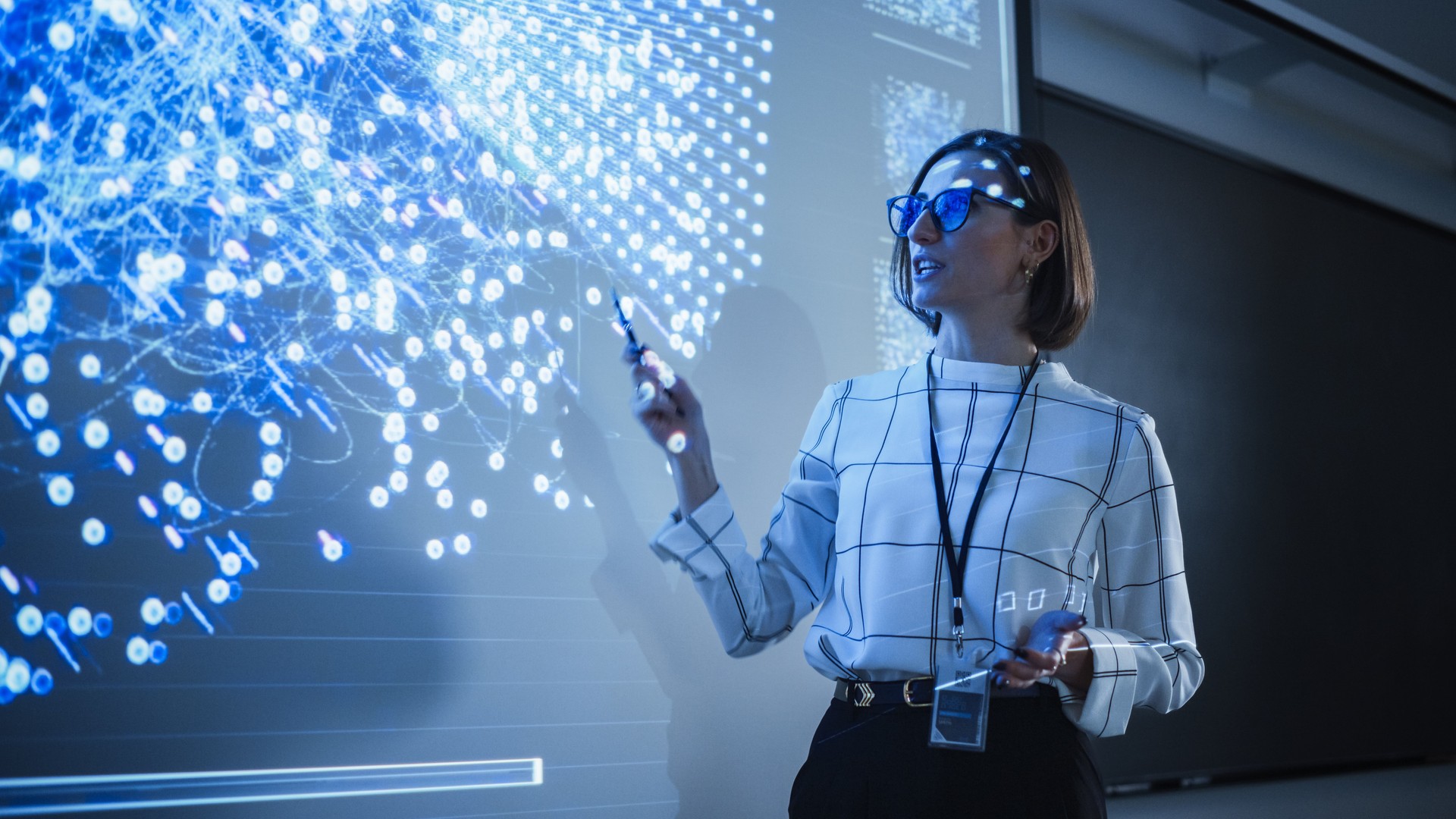 Young University Professor Explaining a Web Development Project to a Group of Students in a Dark Auditorium. Female Teacher Showing a Back-End Programmers Code on a Big Projector Screen